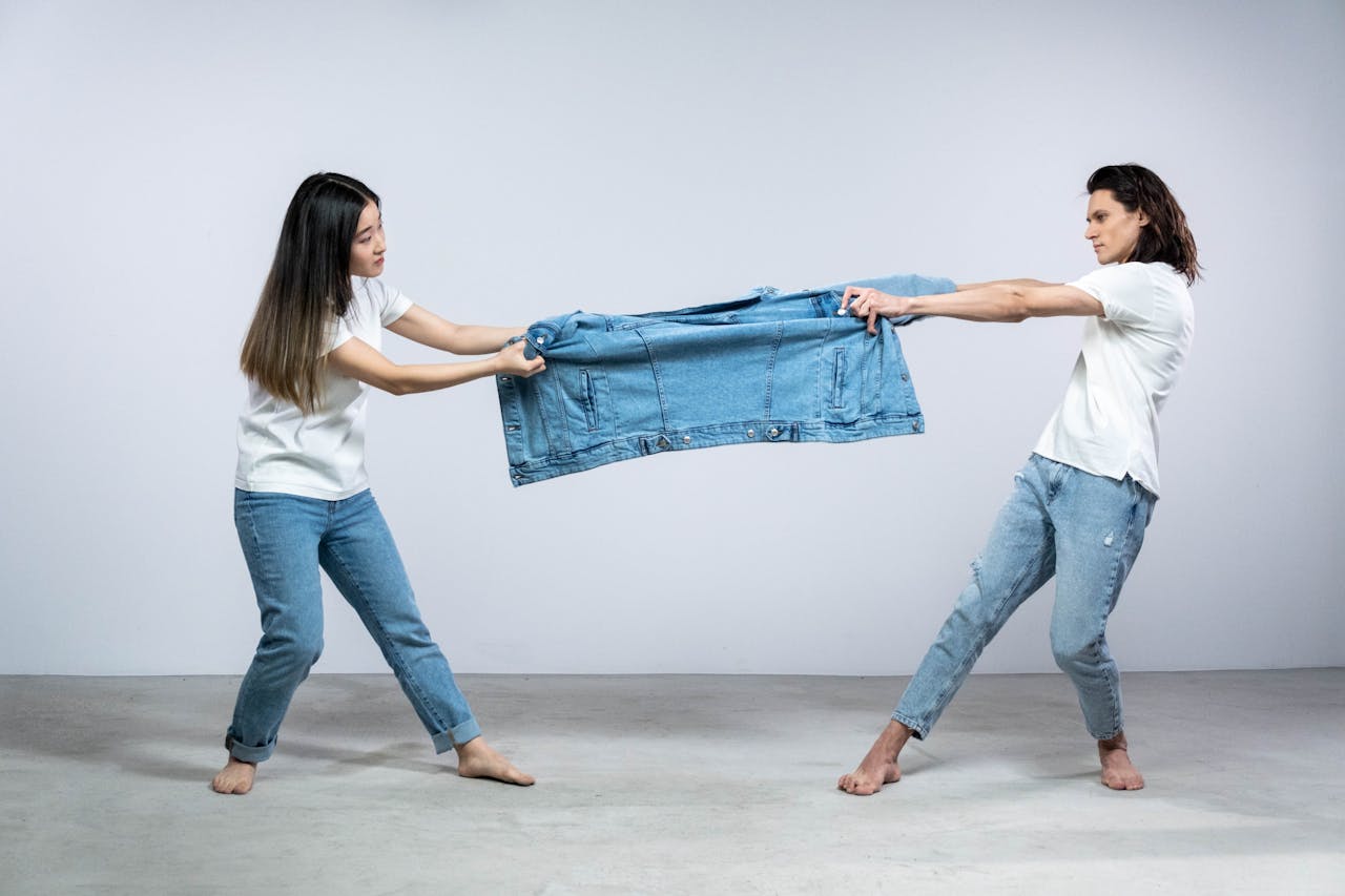Two adults pulling on a denim jacket in a playful fashion pose in a studio setting.